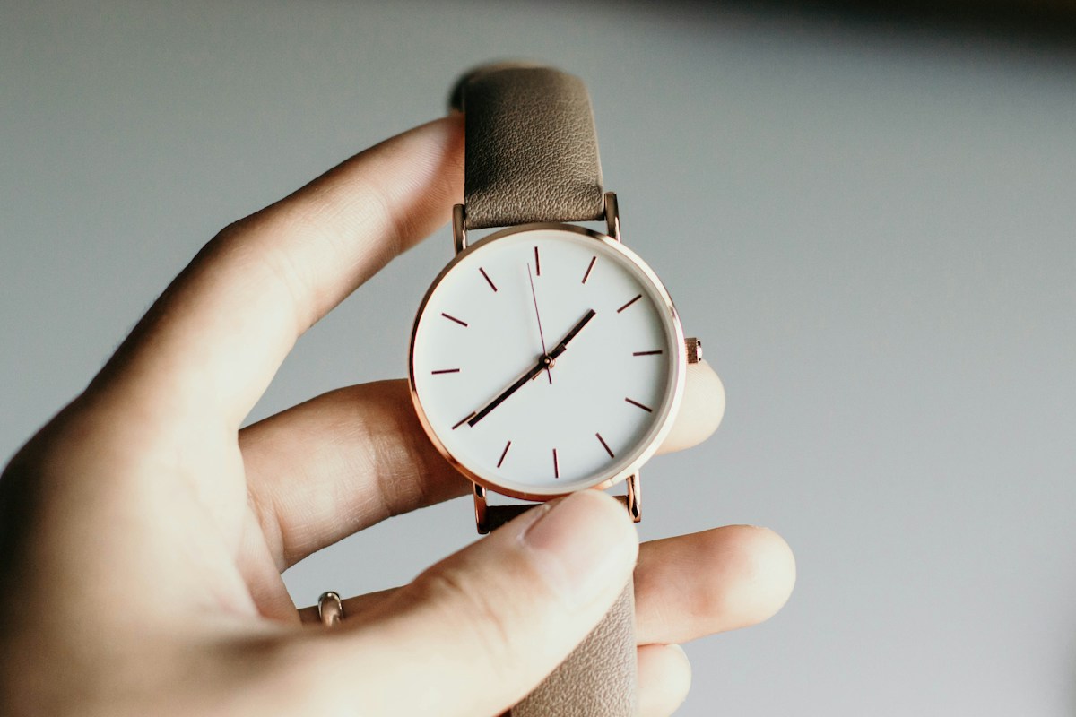 A hand holding a wristwatch with a white dial, rose-gold case and grey leather strap