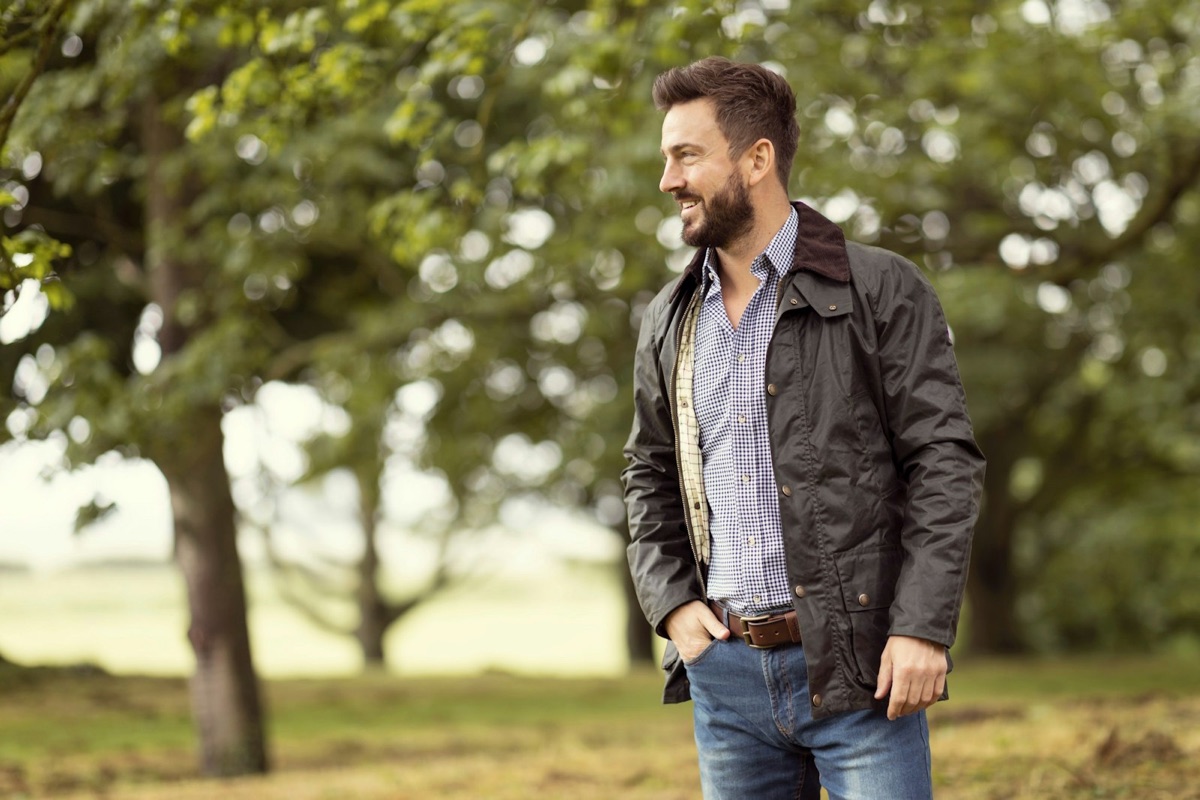A man in a dark green waxed cotton jacket standing among trees in a field